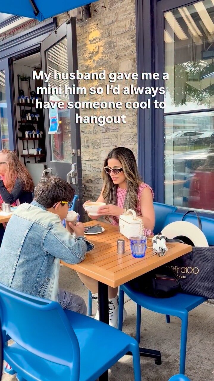 Roopsi Bansal and her son in sunglasses at an outdoor cafe table, both eating from bowl-style cups, with on-screen text reading my husband gave me a mini him so I'd always have someone cool to hangout with