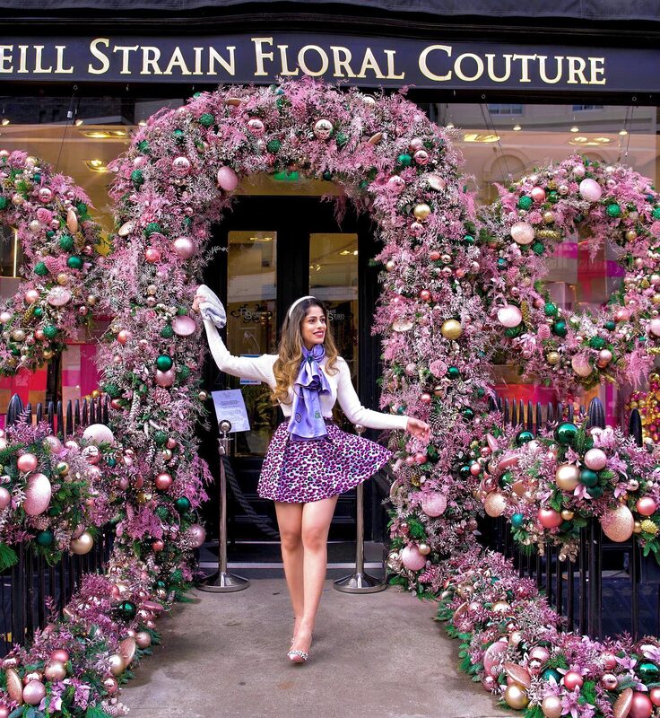 Roopsi Bansal in a white sweater, purple silk scarf and leopard-print mini skirt, walking through a pink floral Christmas archway at Neill Strain Floral Couture
