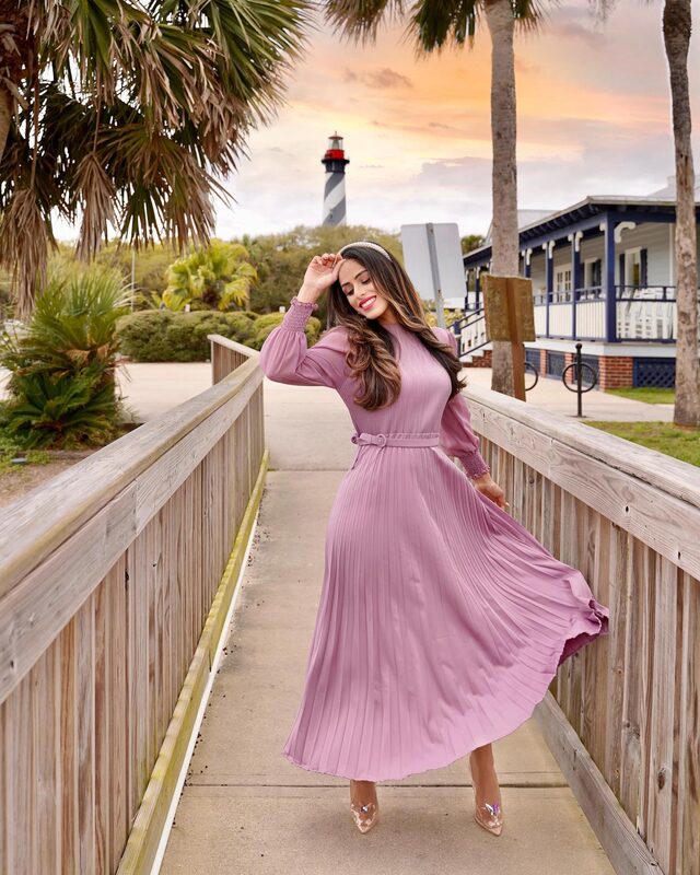 Roopsi Bansal twirling in a mauve pleated long-sleeve maxi dress on a wooden boardwalk, the St. Augustine Lighthouse visible at sunset behind her