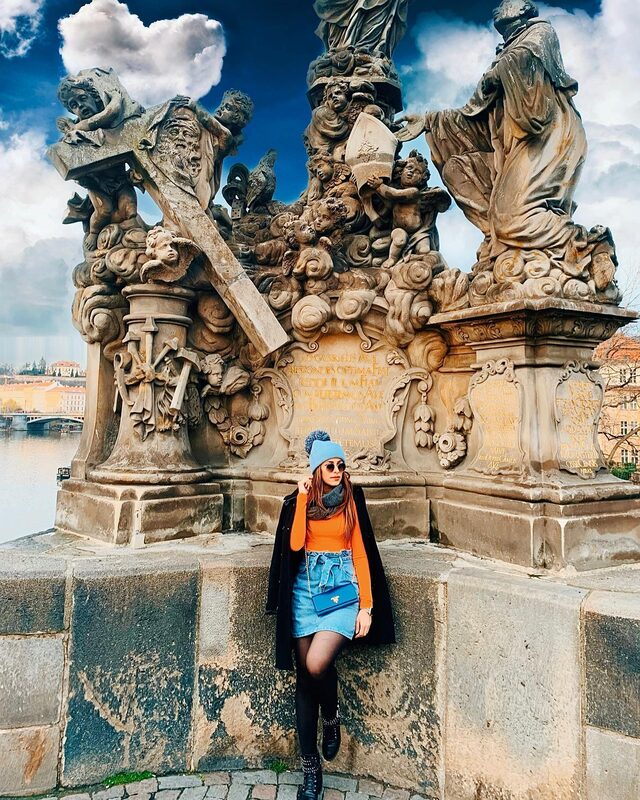 Leaning against the ornate stone base of the Bridge of Saints statue on Prague's Charles Bridge, wearing a black coat, orange sweater, blue pom-pom beanie, and denim skirt, with the river visible behind