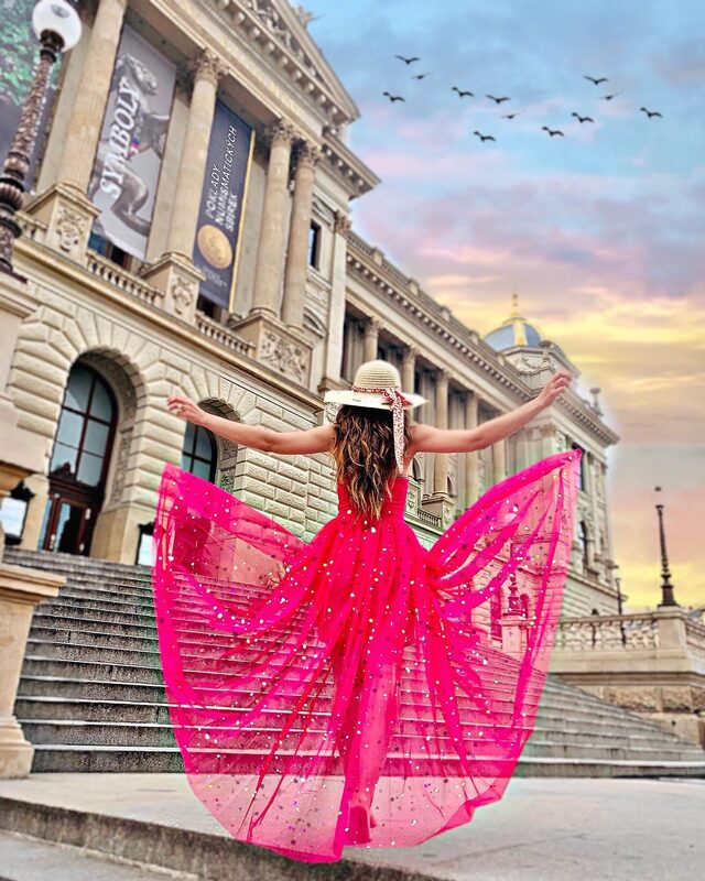Roopsi Bansal in a hot-pink sequined tulle gown and straw hat, arms outstretched on the steps of Prague's National Museum