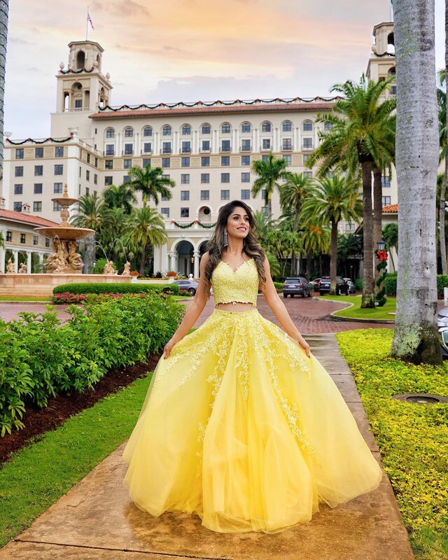 Roopsi Bansal in a yellow lace two-piece ball gown, holding out the tulle skirt, standing on a path in front of The Breakers Palm Beach decorated with holiday garland