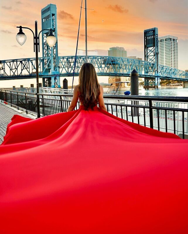 Roopsi Bansal from behind in a red flying dress with a wide train, facing the Main Street Bridge in Jacksonville, Florida at sunset