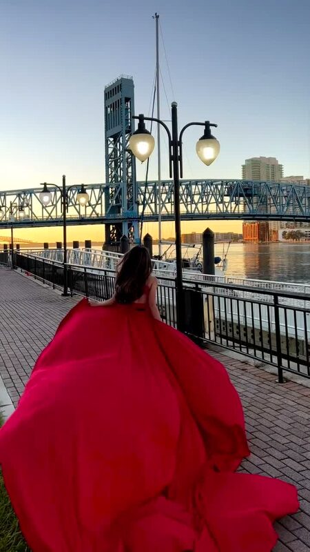 Roopsi Bansal from behind in a flowing red gown on the riverwalk at the Main Street Bridge in Jacksonville, Florida, at sunset