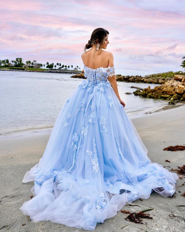 Roopsi Bansal from behind in a sky-blue off-shoulder ball gown covered in floral lace appliqués, walking along the sandy shore at Dubois Park, Florida, beside a rocky jetty