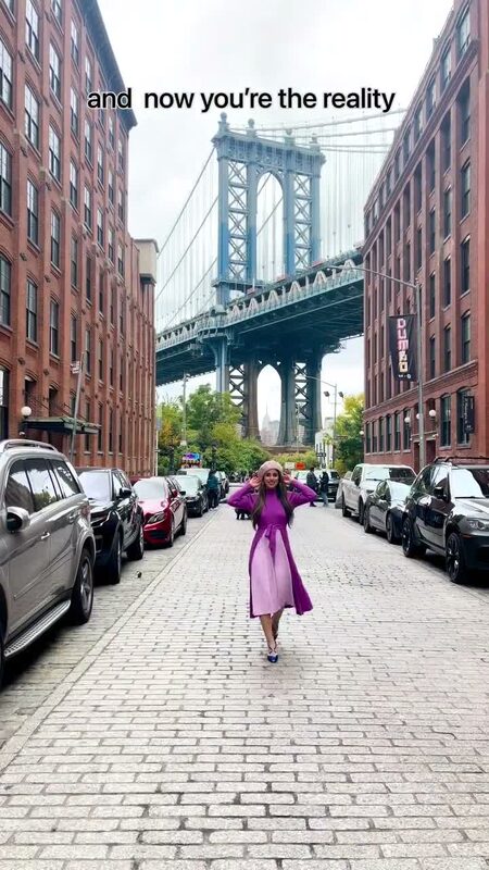 Roopsi Bansal in a purple wrap dress standing on a cobblestone street in Dumbo with the Manhattan Bridge behind her