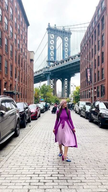 Roopsi Bansal in a purple wrap dress walking down a cobblestone street in Dumbo, Brooklyn, framed by the Manhattan Bridge
