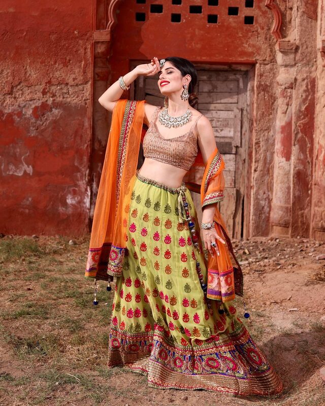 Roopsi Bansal in a pistachio green and orange embroidered lehenga with a statement necklace, posing against a weathered terracotta-red wall for Eid