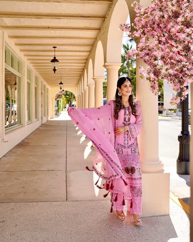 Roopsi Bansal in a pink embroidered salwar suit with a tasseled dupatta from Khas, leaning against a colonnade pillar beneath a blossoming pink tree in West Palm Beach