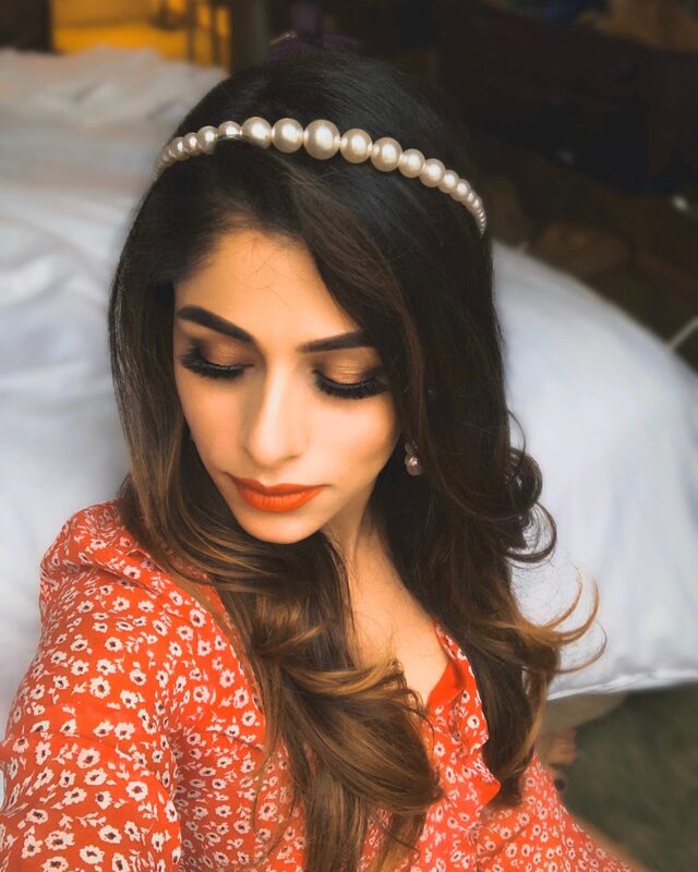 Close-up portrait in a red floral wrap dress and pearl headband, warm makeup and a bold orange-red lip, sitting on a bed