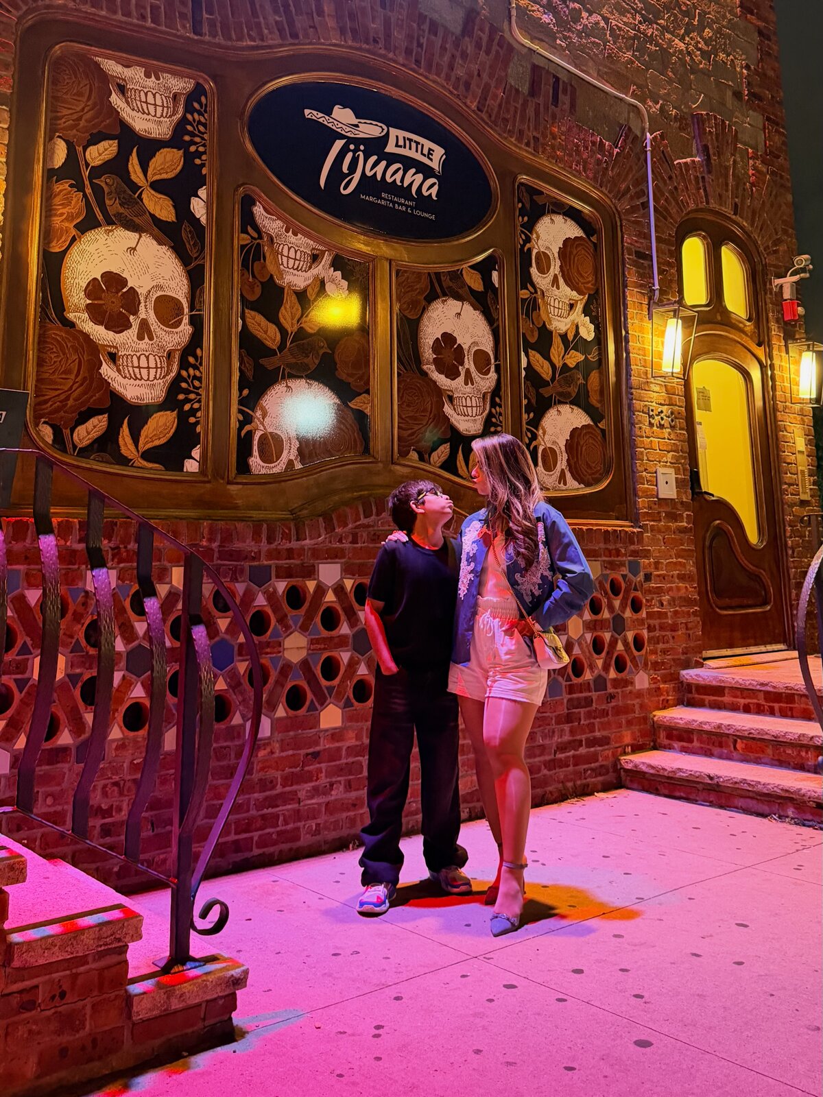 Roopsi Bansal and her son posing in front of a wall of painted skull murals lit in pink