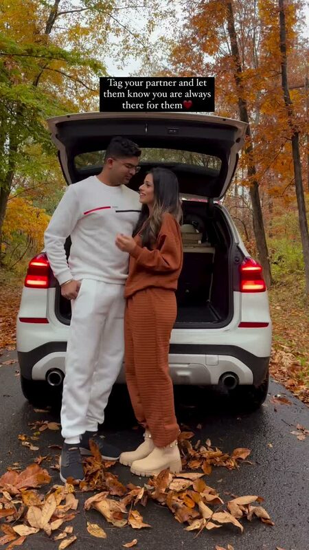 Roopsi Bansal and her husband in matching cozy sweatsuits by the open trunk of their car on a leaf-covered wooded lane