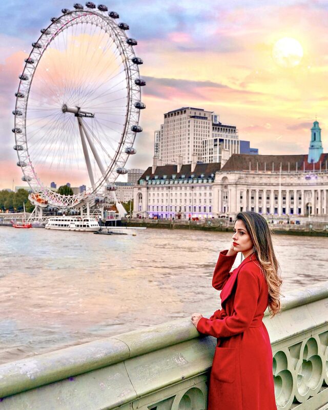 Roopsi Bansal in a red coat, leaning on a bridge railing with the London Eye and the Thames in the background at sunset