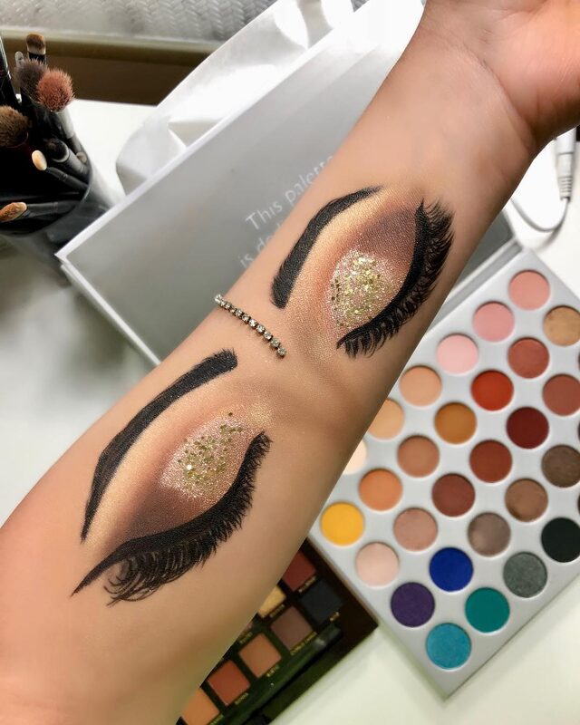 A warm glittery gold eyeshadow look swatched onto a forearm, with makeup brushes and an eyeshadow palette laid out alongside