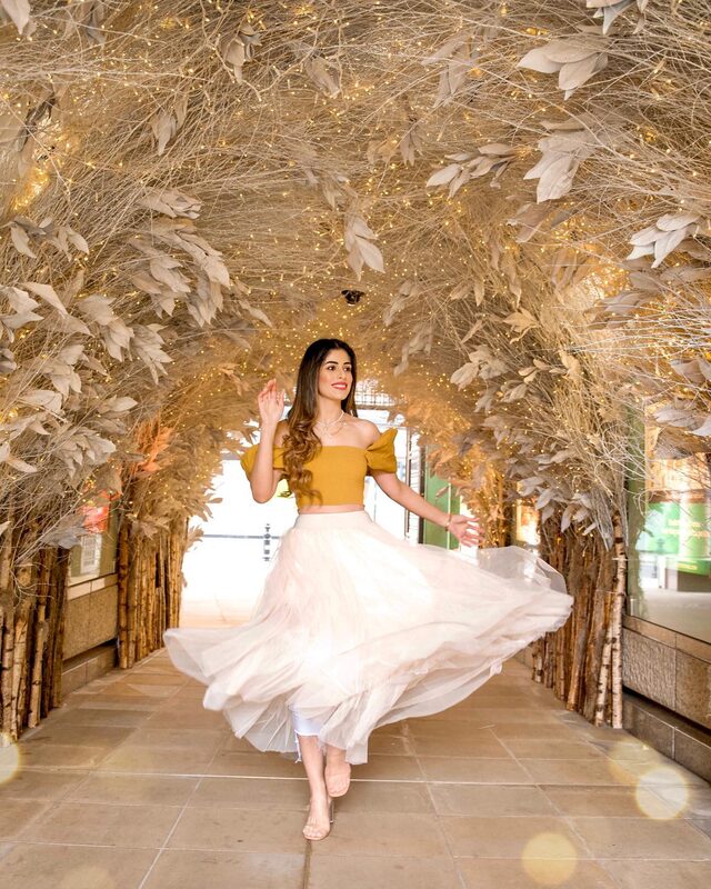 Roopsi Bansal twirling in a mustard off-shoulder crop top and a blush tulle skirt inside a fairy-lit tunnel of woven branches at Duke of York Square, London