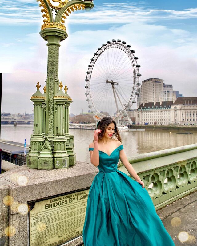 Roopsi Bansal in a teal off-shoulder ball gown, standing on Westminster Bridge beside an ornate lamppost with the London Eye and the Thames behind her