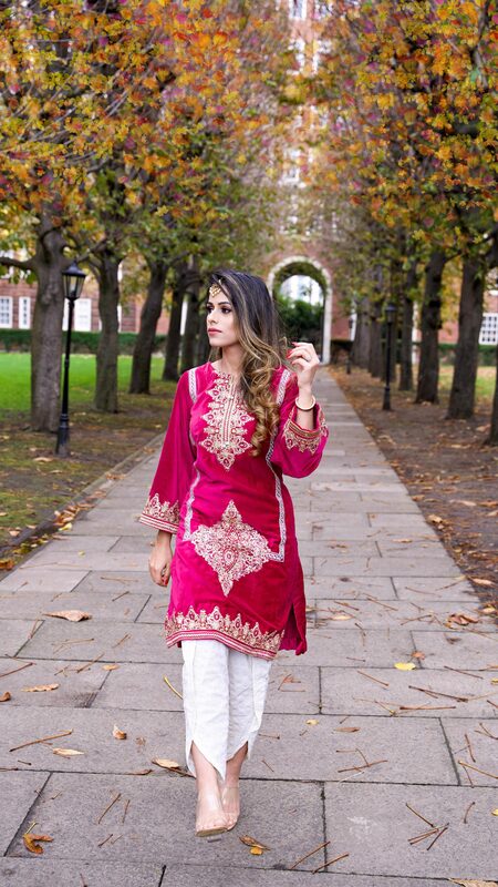 Roopsi Bansal in a magenta velvet embroidered kurta with white dhoti pants, walking a tree-lined path under a brick archway for Diwali