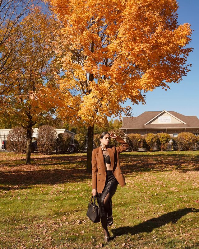 Roopsi Bansal in a rust double-breasted blazer, black lace bralette, and leather skirt standing under a maple tree with brilliant orange fall leaves