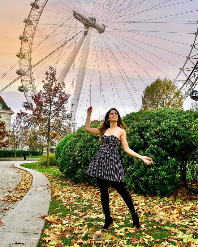 Roopsi Bansal in a strapless black and white polka dot dress, mid-twirl in fallen autumn leaves beside a hedge with the London Eye turning behind her