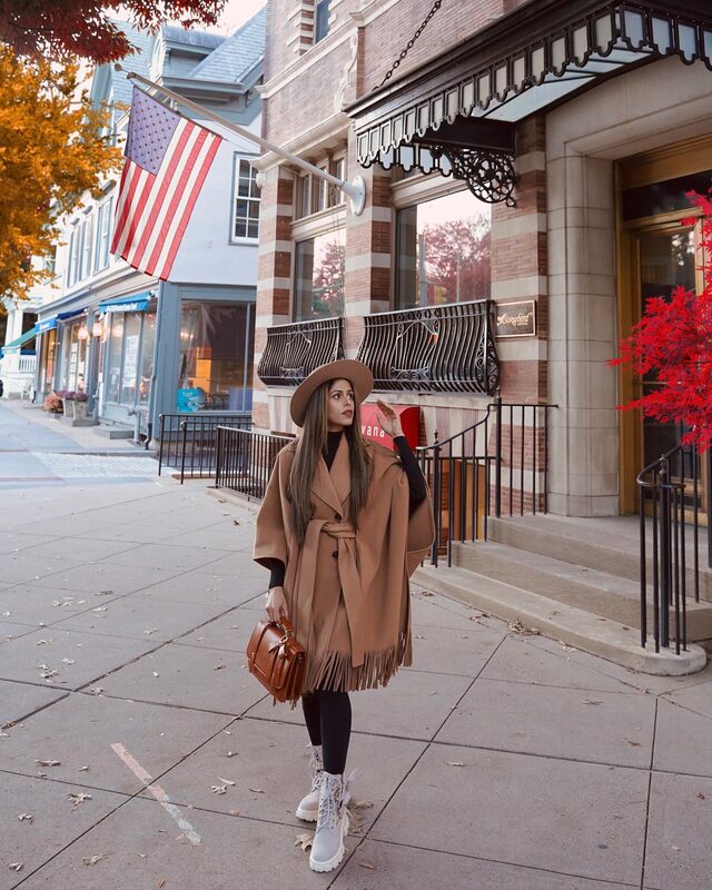 Roopsi Bansal in a camel fringe poncho coat and wide-brim hat walking a small-town main street lined with American flags and fall foliage