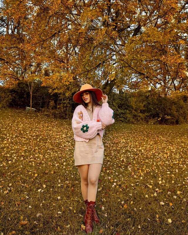 Roopsi Bansal in a pink hand-knit chunky cardigan with flower appliques, a wide floppy hat, and a tan mini skirt, standing in a field covered in fallen golden leaves
