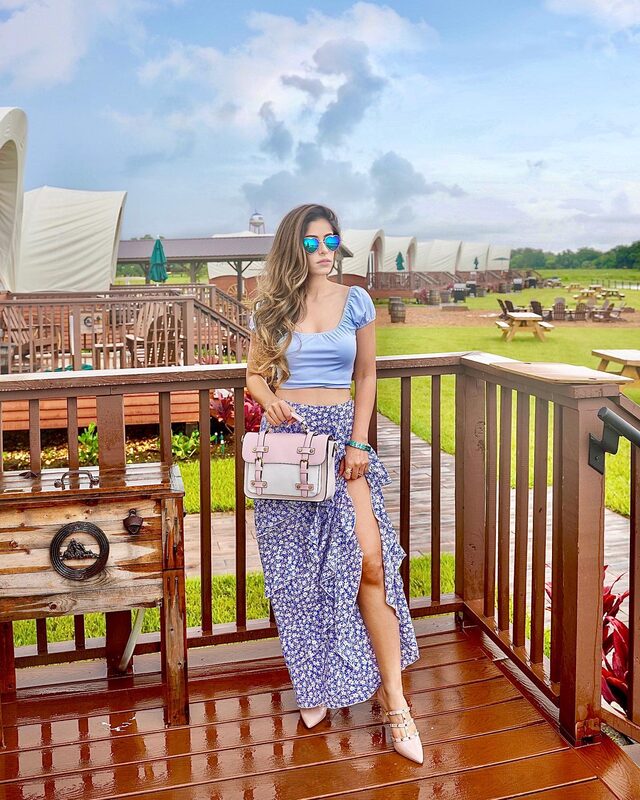 Roopsi Bansal in a light blue crop top and blue floral wrap skirt, mirrored sunglasses, holding a pink handbag on a wooden deck at Westgate River Ranch with glamping tents behind her