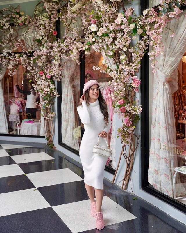 Roopsi Bansal in a white pearl-decorated turtleneck bodycon dress and pink beret, standing under an archway of pink cherry blossoms outside a storefront