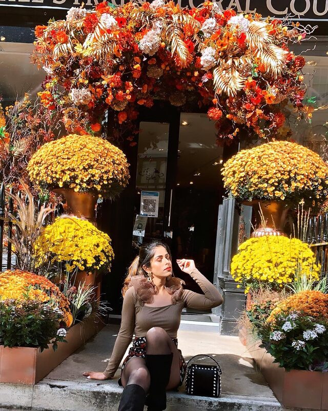 Roopsi Bansal in a brown ruffle-sleeve top and floral mini skirt with black over-the-knee boots, sitting among orange and yellow chrysanthemum planters outside a florist