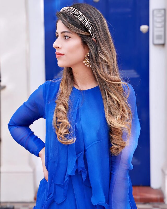 Roopsi Bansal in a royal blue sheer-sleeve outfit with a jeweled headband and drop earrings, standing in front of a blue door