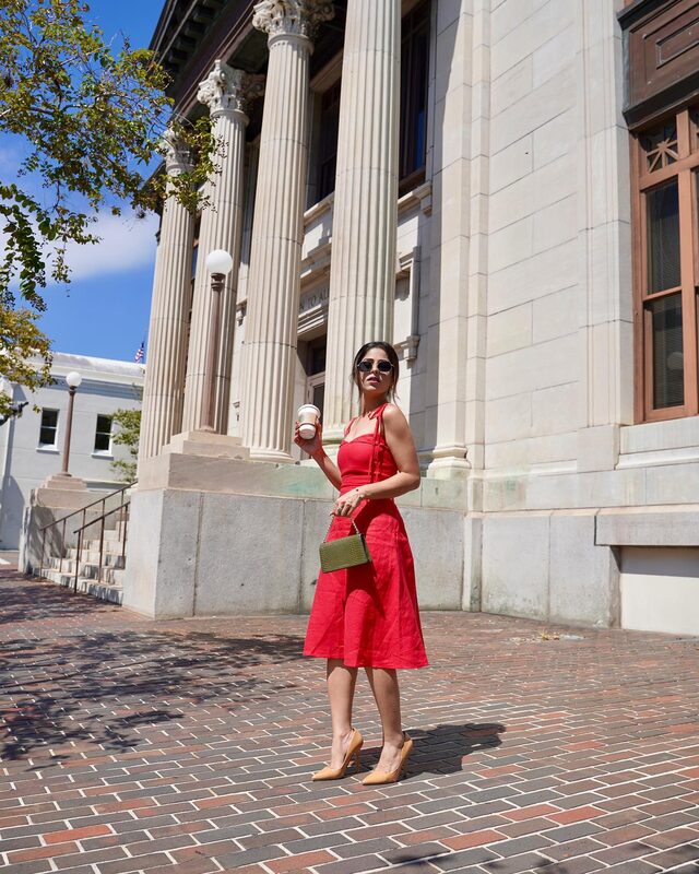 Roopsi Bansal in a red midi dress, holding a coffee cup and a small olive green bag, standing in front of tall stone columns in Jacksonville, Florida
