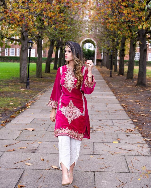Roopsi Bansal in a magenta velvet embroidered kurta with white bottoms and gold jewelry, walking down a tree-lined path