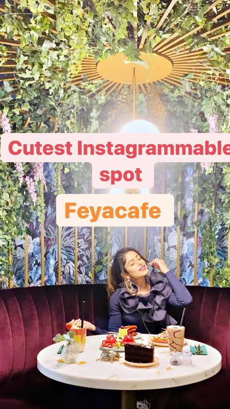Roopsi Bansal in a navy ruffle-collar top, seated at a marble table with a brunch spread including cake, drinks and pastries, under a botanical-domed ceiling