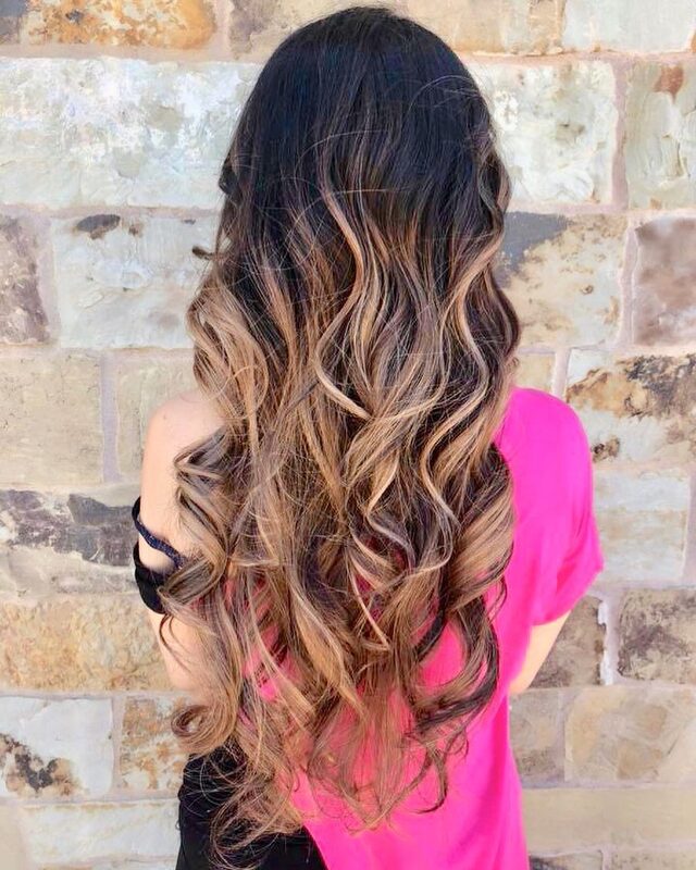 Long balayage hair in soft curls, viewed from behind, against a stone wall, wearing a pink top