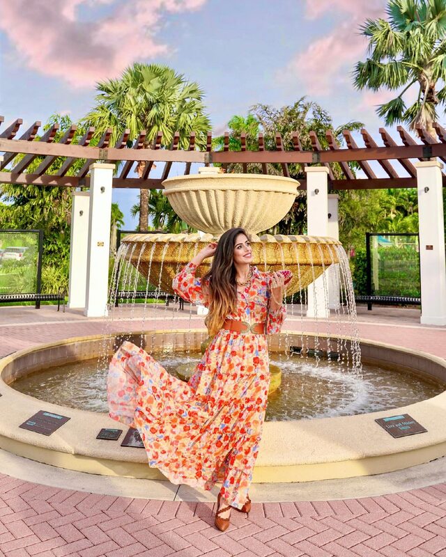 Roopsi Bansal in a colourful floral print maxi dress with a wide brown belt, sitting on the edge of a tiered stone fountain in Wellington, Florida