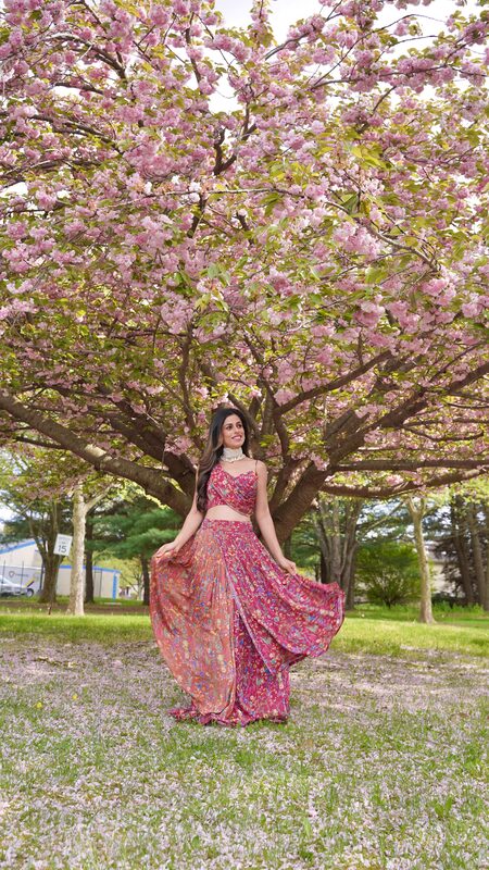 Roopsi Bansal in a floral lehenga set standing under a full-bloom pink cherry blossom tree with petals covering the grass