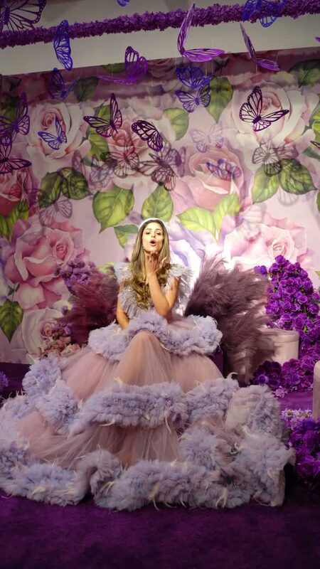 Roopsi Bansal in a blush tulle ball gown with feathered ruffle tiers, seated under hanging paper butterflies in front of a rose-covered wall at The Floral Escape