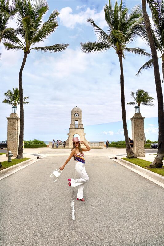 Roopsi Bansal in a colorful bandana-print bandeau top and white flare pants, kicking up a heel on the palm-lined road leading to the Worth Avenue clock tower in West Palm Beach