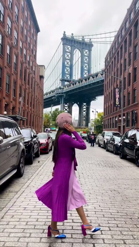 Roopsi Bansal in a magenta pleated dress on a cobblestone DUMBO street, the Manhattan Bridge framed behind her