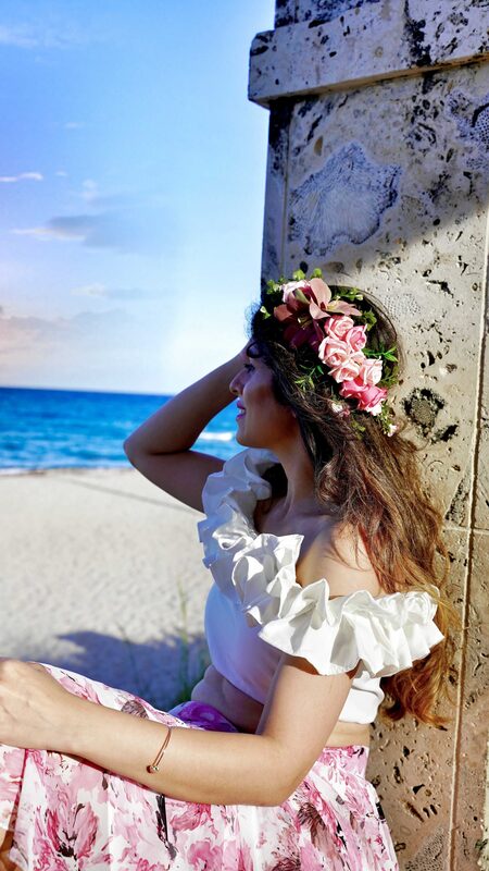 Roopsi Bansal in a pink floral skirt and white ruffle top wearing a flower crown, looking out at the ocean beside a coral-stone wall