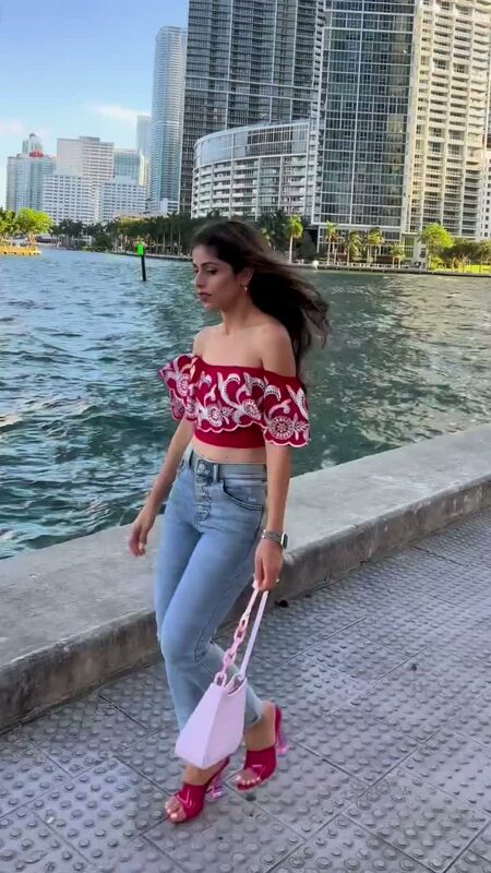 Roopsi Bansal in a red embroidered off-shoulder ruffle crop top and ripped skinny jeans from Express, carrying a pink handbag, walking along the water at Bayfront Park with the Miami skyline behind her