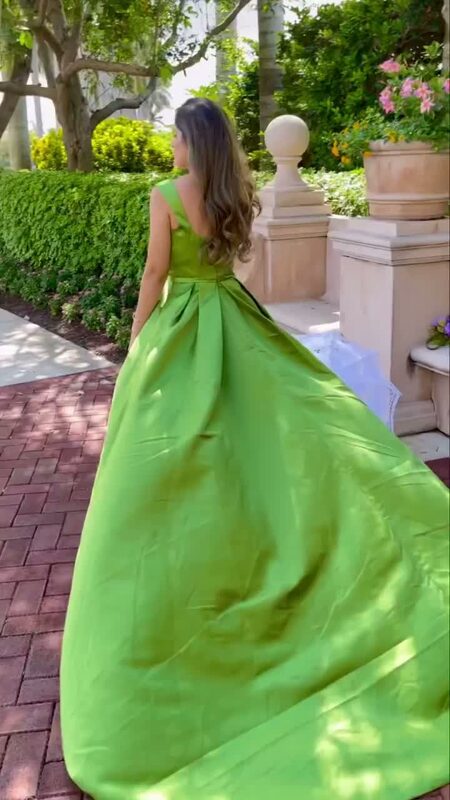 Roopsi Bansal from behind in a bright green satin ball gown with a full train, walking down a brick garden path lined with hedges and stone urns
