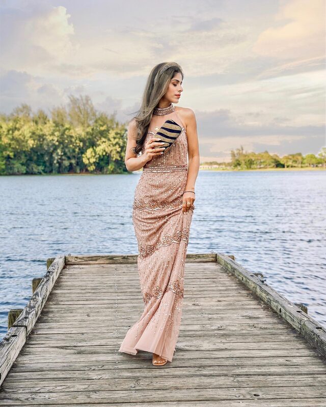 Roopsi Bansal in a hand-beaded rose gold illusion halter gown from Adrianna Papell, holding a striped beaded clutch from Jaipur Rose, standing at the end of a wooden dock over the water