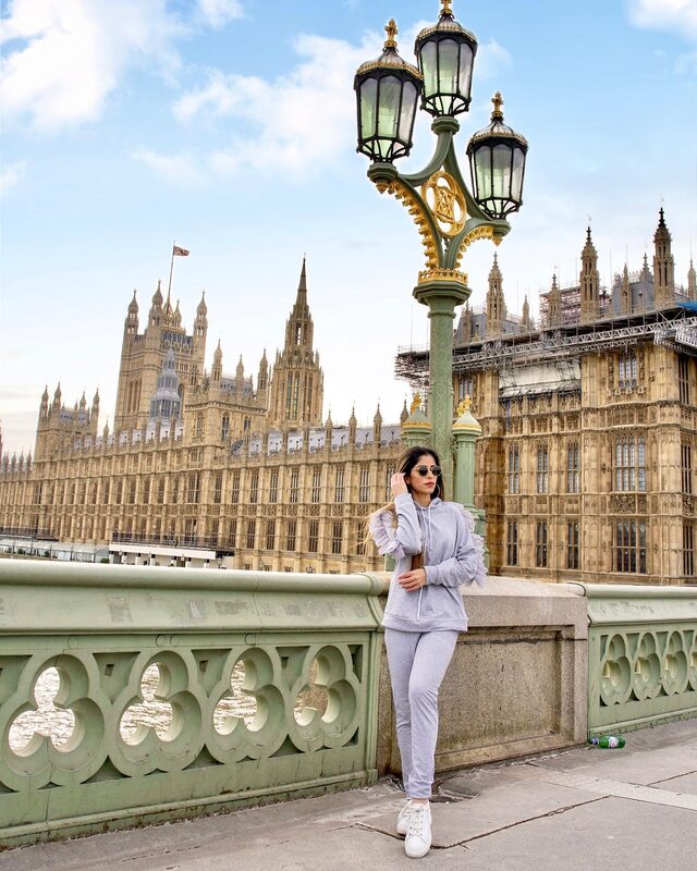 Roopsi Bansal in a grey ruffle-trim loungewear set and sunglasses, leaning against Westminster Bridge under an ornate lamppost with the Houses of Parliament behind her
