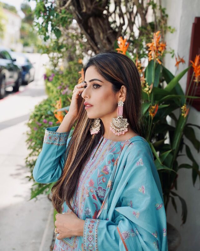 Roopsi Bansal in profile wearing a teal Kani-print ethnic set and statement gold-and-pink chandelier earrings against a backdrop of tropical flowers