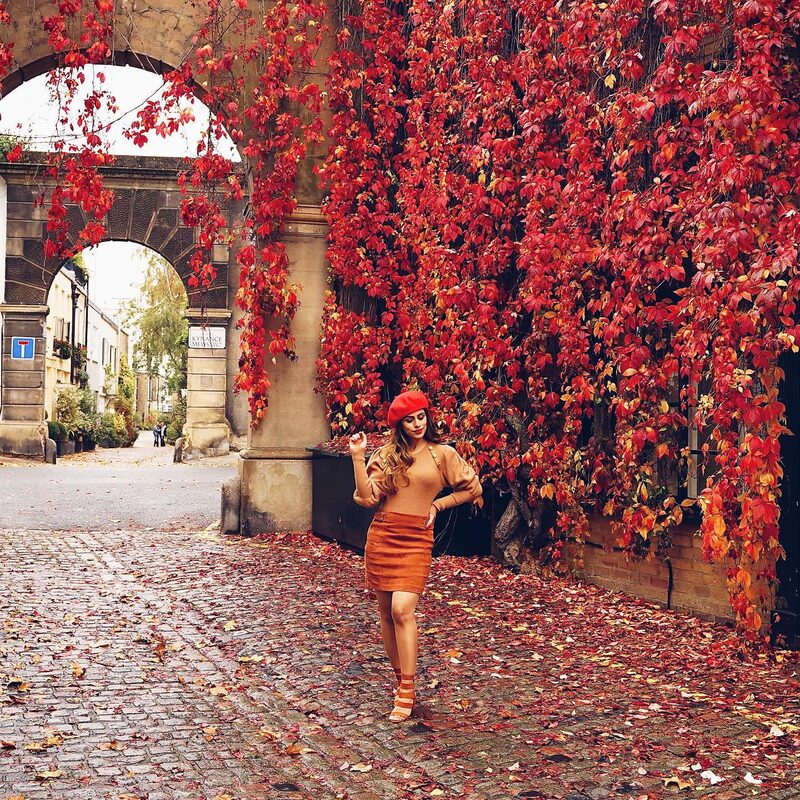 Roopsi Bansal in a rust-colored puff-sleeve sweater, matching skirt and red beret, standing beneath the red autumn ivy at Kynance Mews
