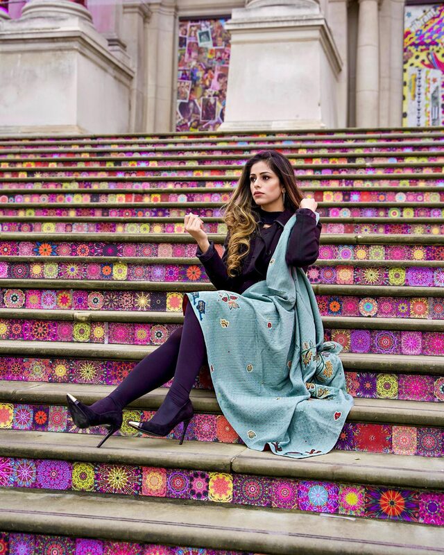 Roopsi Bansal in a black outfit with a dusty-teal embroidered wrap, sitting on Tate Britain's colorful kaleidoscope-patterned steps