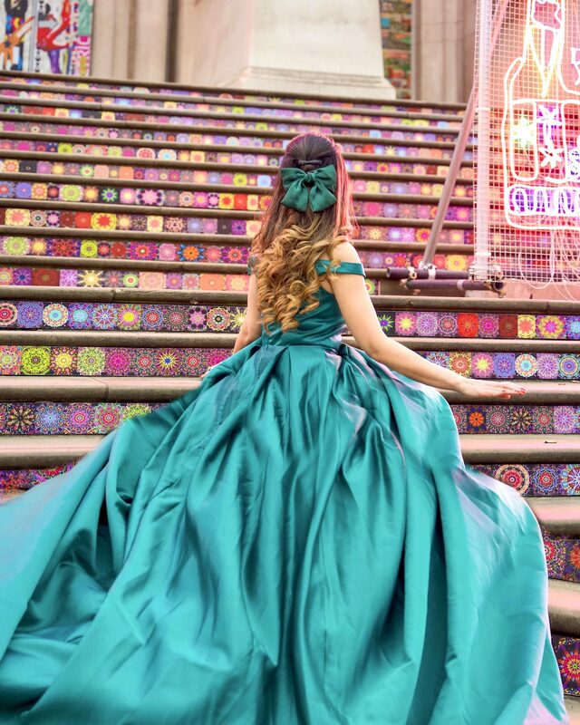 Roopsi Bansal from behind in a teal off-shoulder ball gown with a full satin skirt and a teal bow in her hair, walking up colourful mosaic-patterned steps