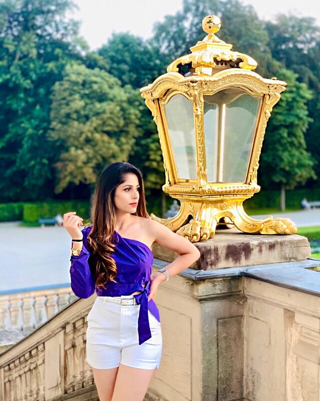 Roopsi Bansal in a purple one-shoulder top and white shorts, posing beside an ornate gold lamppost in a Munich garden