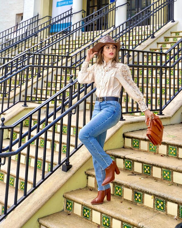 Roopsi Bansal in a white lace blouse, straw cowboy hat, and jeans, descending colorful tiled steps at the Harriet Himmel Theater in Rosemary Square, holding a rust-colored fringed bag