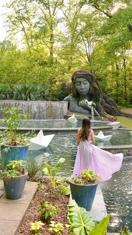 Roopsi Bansal in a lilac wrap dress from SHEIN, standing before the Earth Goddess mosaiculture sculpture and reflecting pool at Atlanta Botanical Garden